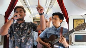 Two musicians are performing inside an airplane. The person on the left is singing into a microphone and has one hand raised, while the person on the right is playing an acoustic guitar. Both are standing in the aisle of the plane, and there are passengers seated behind them. The interior of the airplane is visible, including overhead compartments and a sign that reads "SKYTRAX."